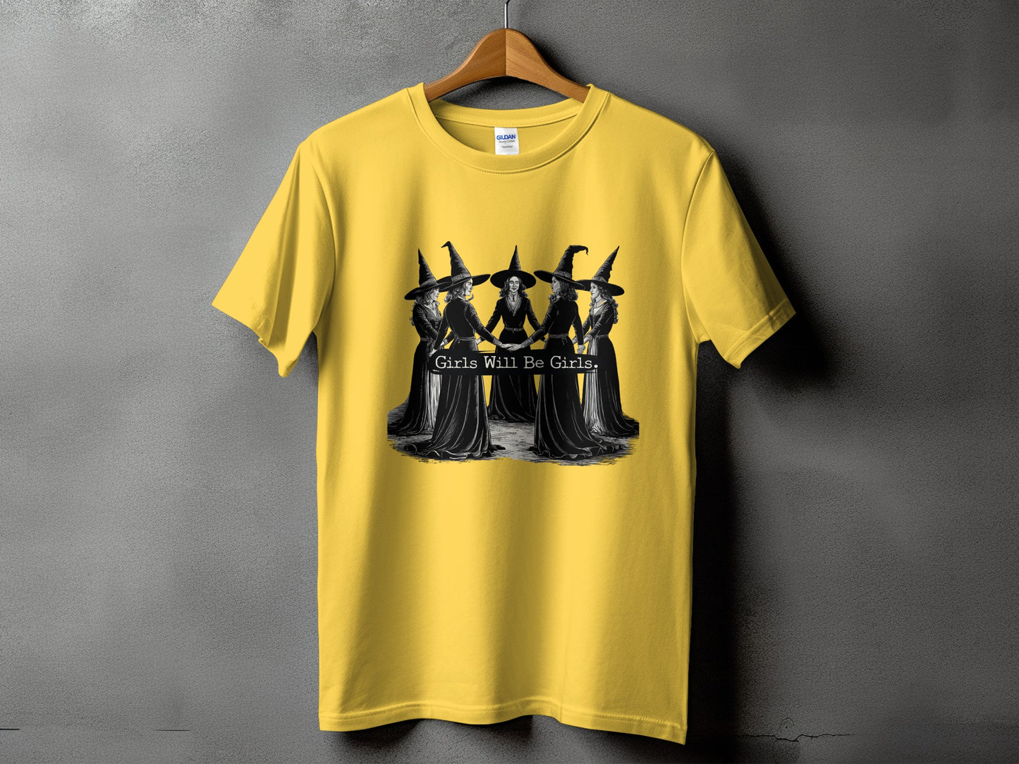 Yellow t-shirt with black graphic of witches and text "Girls Will Be Girls."
