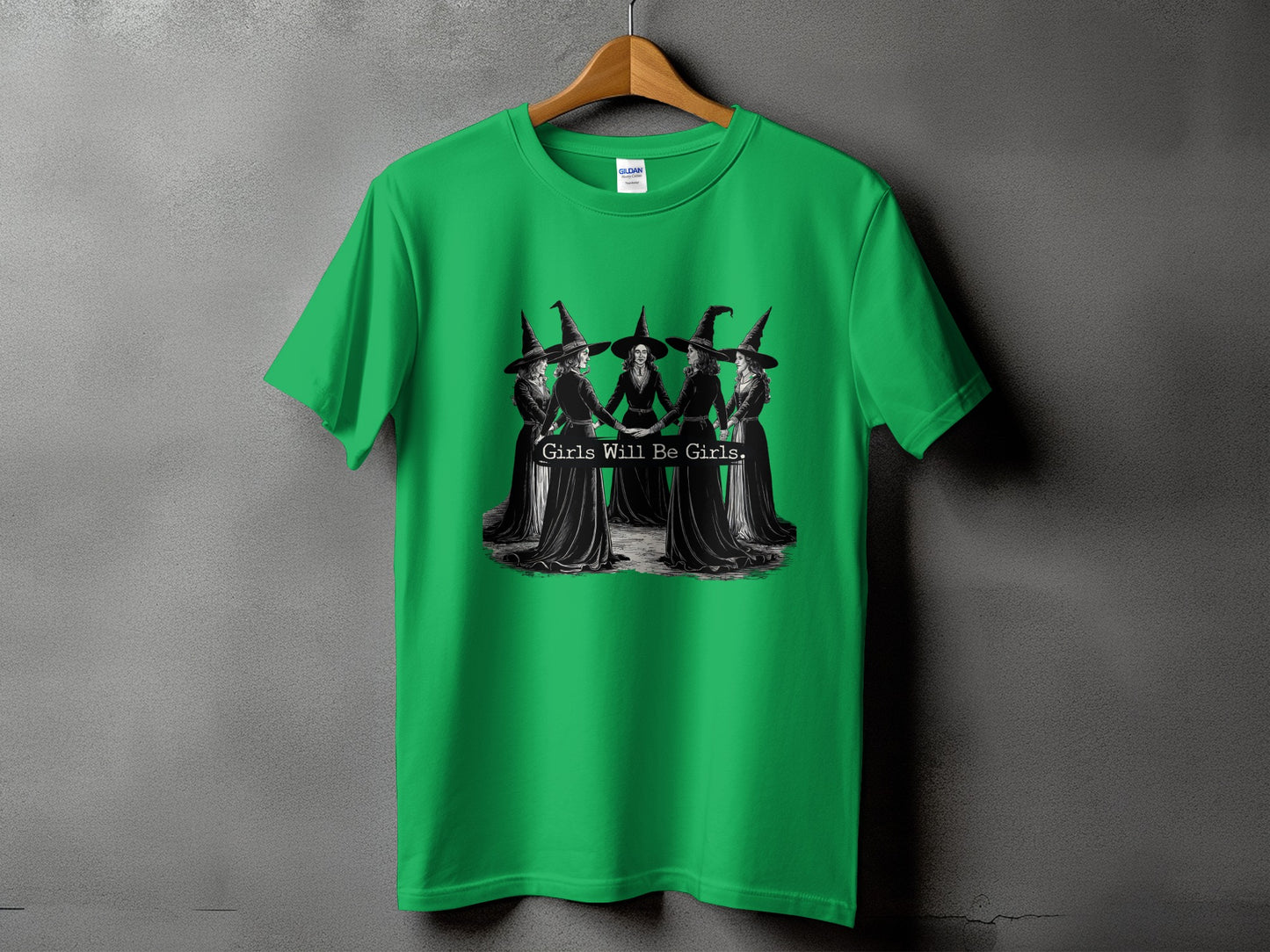 Green t-shirt with a graphic of witches holding hands and the text "Girls Will Be Girls".