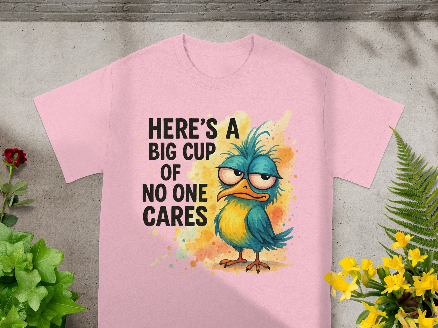 Pink t-shirt with a grumpy blue and yellow bird and text "HERE'S A BIG CUP OF NO ONE CARES".