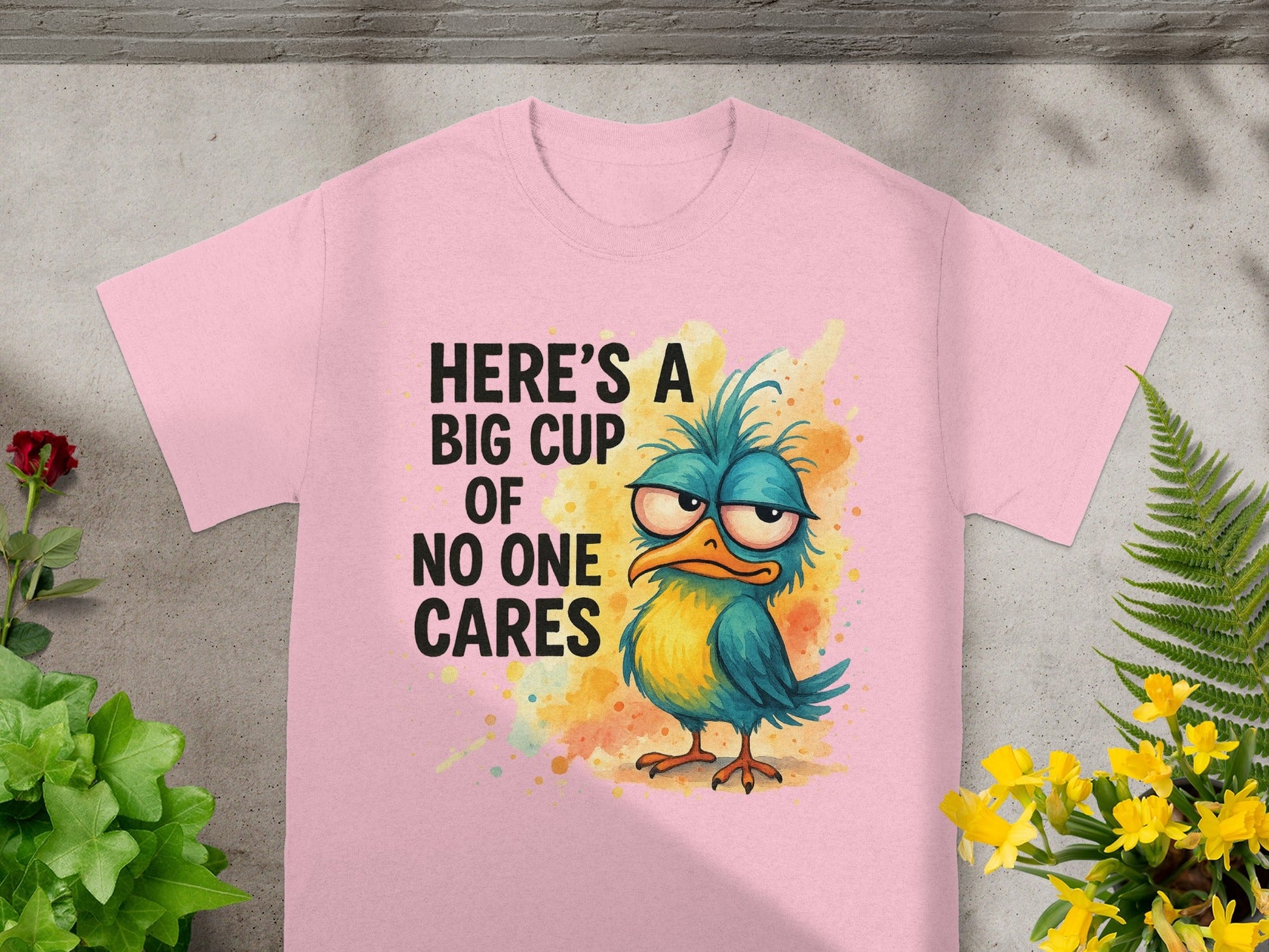 Pink t-shirt with a grumpy blue and yellow bird and text "HERE'S A BIG CUP OF NO ONE CARES".
