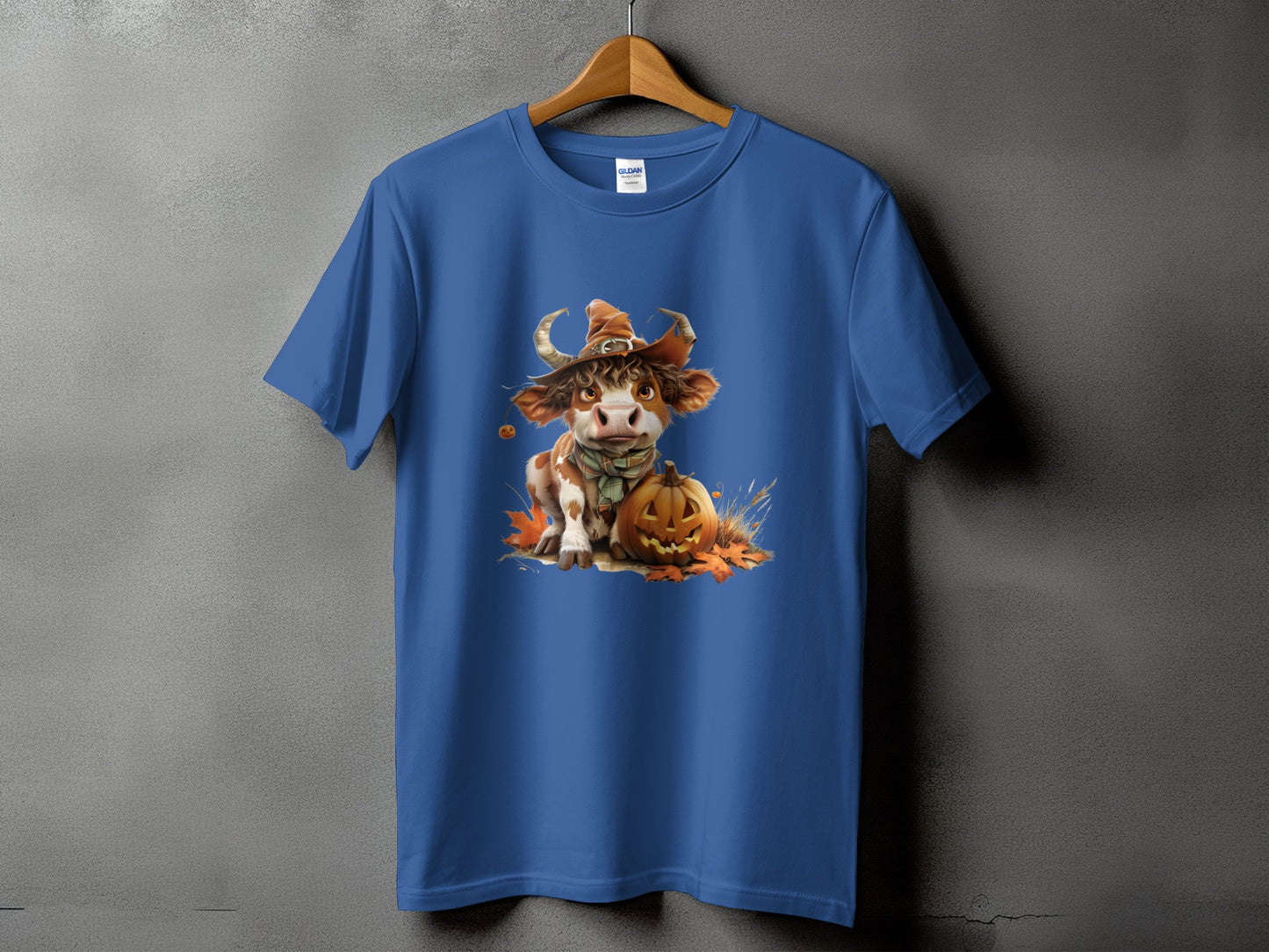 Blue t-shirt with a cartoon cow wearing a witch hat, sitting by a pumpkin.