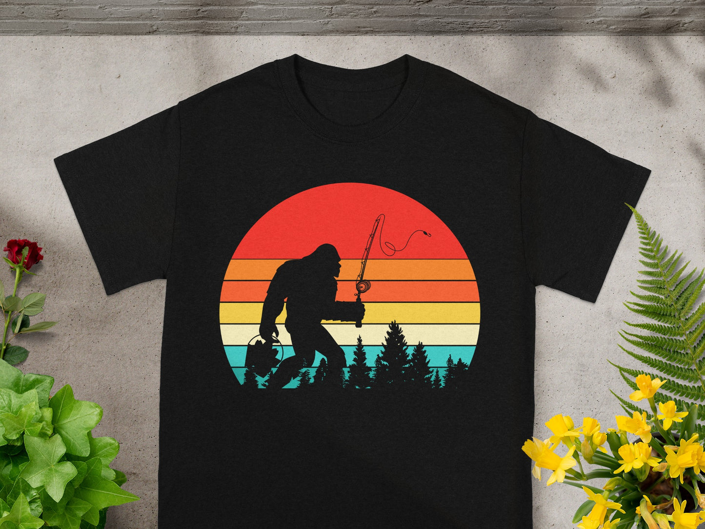 Bigfoot fishing with a sunset and forest graphic on a black t-shirt.