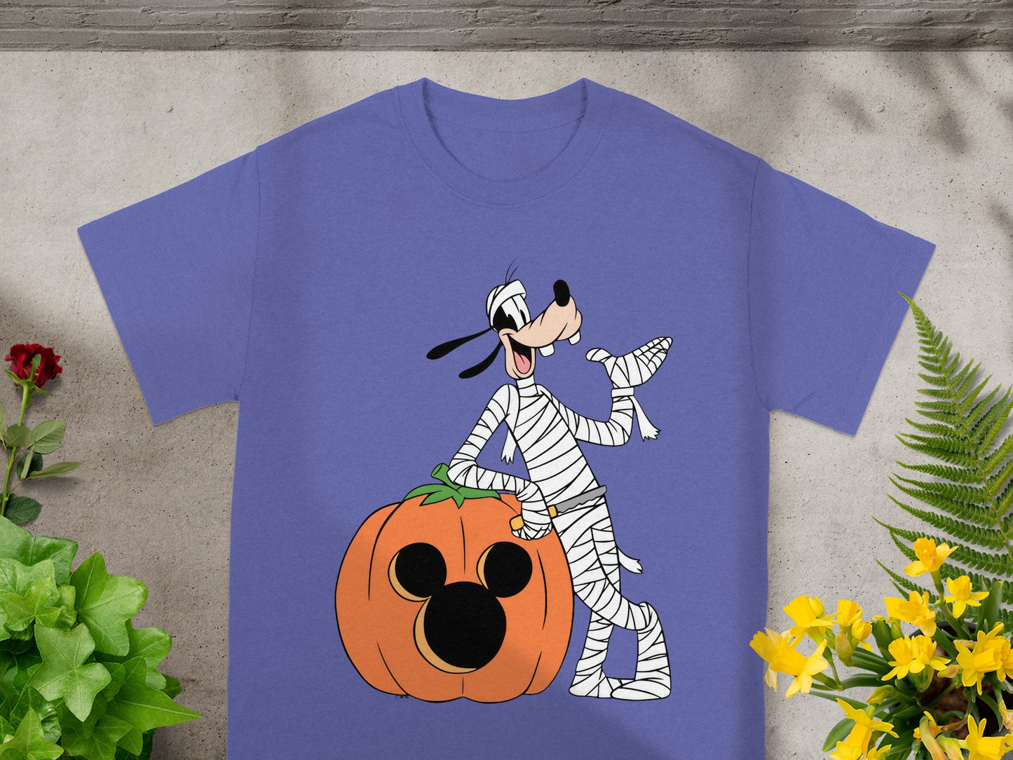 Goofy dressed as a mummy leans on a pumpkin with Mickey Mouse ears on a purple t-shirt.