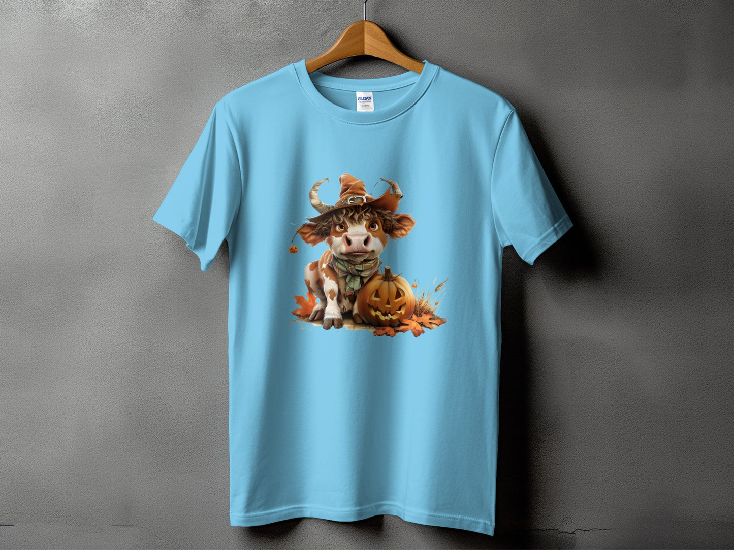 Light blue t-shirt with a cartoon cow wearing a witch hat and scarf, sitting by a pumpkin.