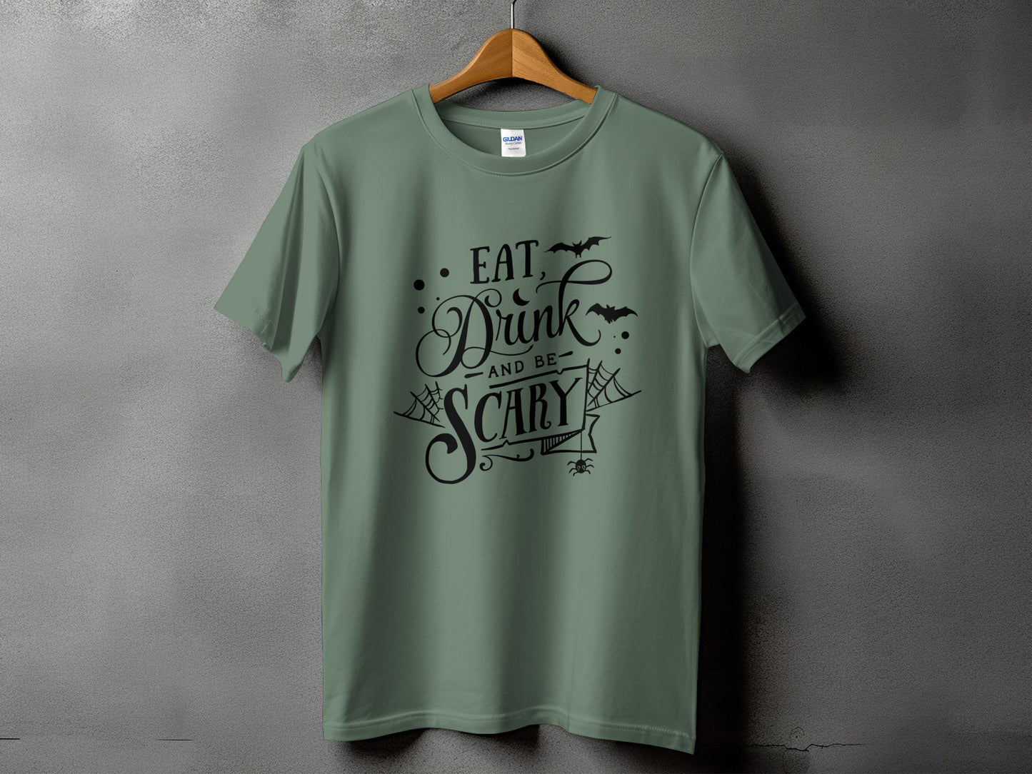 Sage green t-shirt with black text "Eat, Drink, and Be Scary" and Halloween graphics.