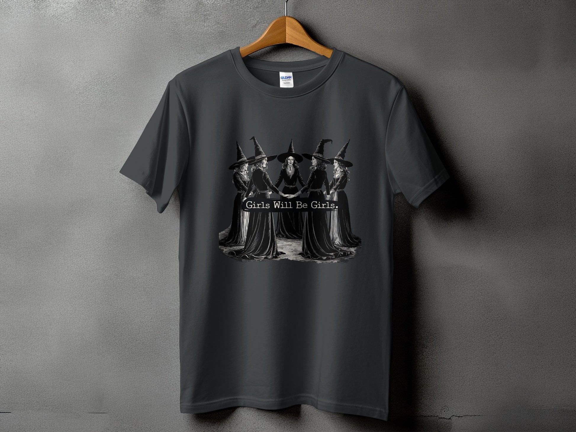 Dark t-shirt with a black and white graphic of women in witch hats holding hands and the text "Girls Will Be Girls."