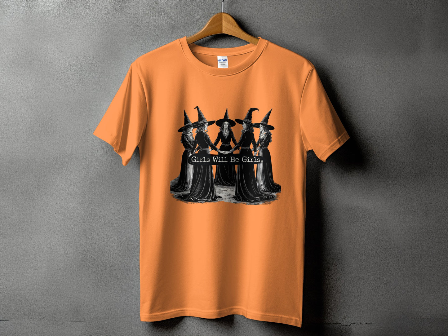 Orange t-shirt with a graphic of witches holding hands and the text "Girls Will Be Girls."