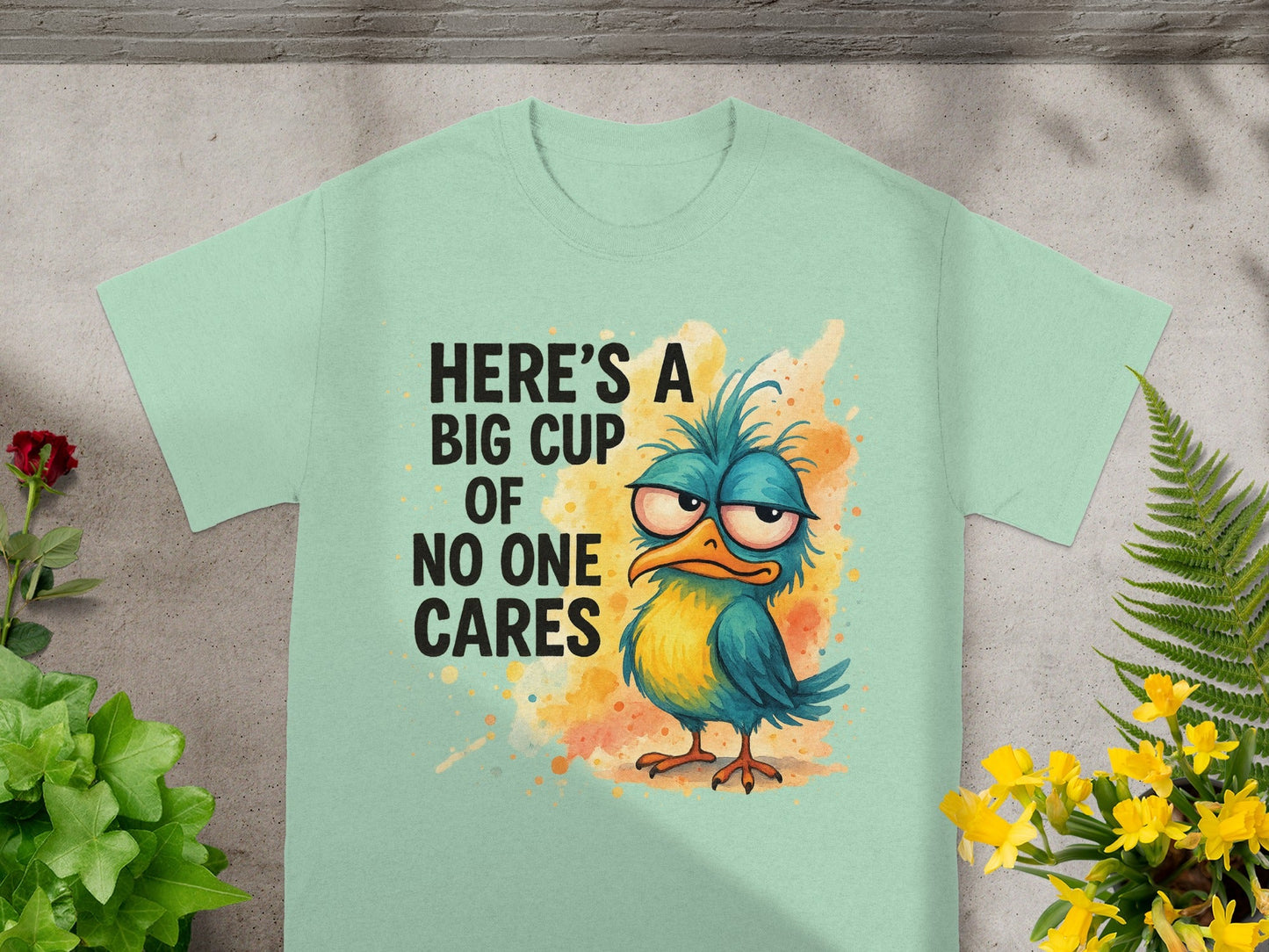 Light blue t-shirt with a grumpy blue and yellow bird and text that reads "HERE'S A BIG CUP OF NO ONE CARES".