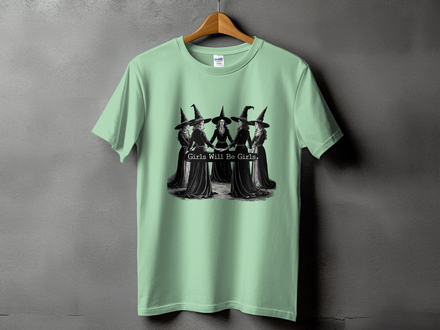 Light green t-shirt with black graphic of five witches holding hands with text "Girls Will Be Girls."