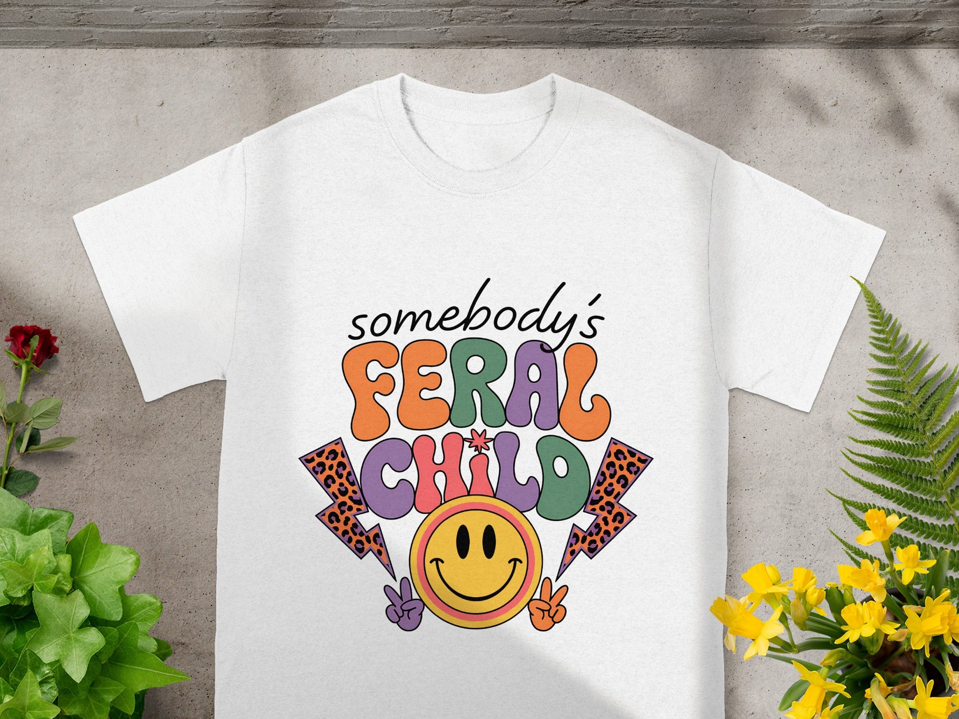 White t-shirt with "somebody's FERAL CHILD" graphic featuring smiley face and lightning bolts.