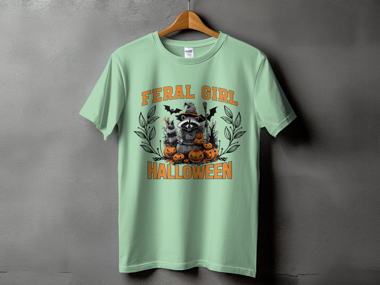 "Feral Girl Halloween" graphic with raccoon, pumpkins, bats, and witch's hat on a mint t-shirt.