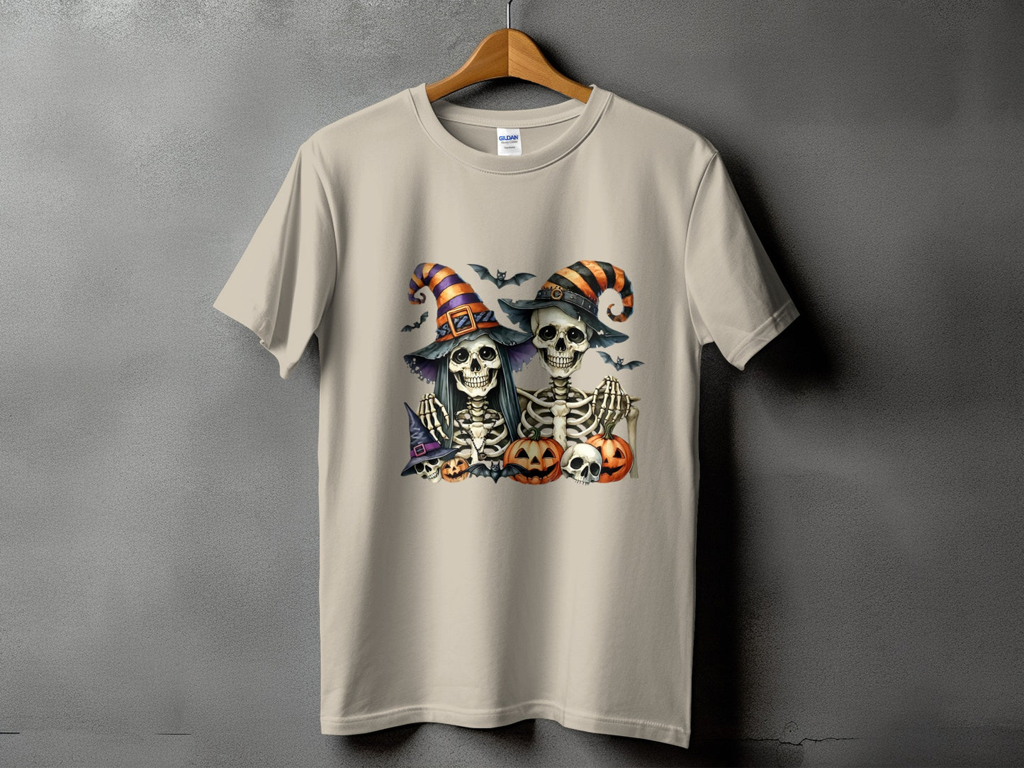 Two skeletons in witch hats with pumpkins and bats on a tan t-shirt.