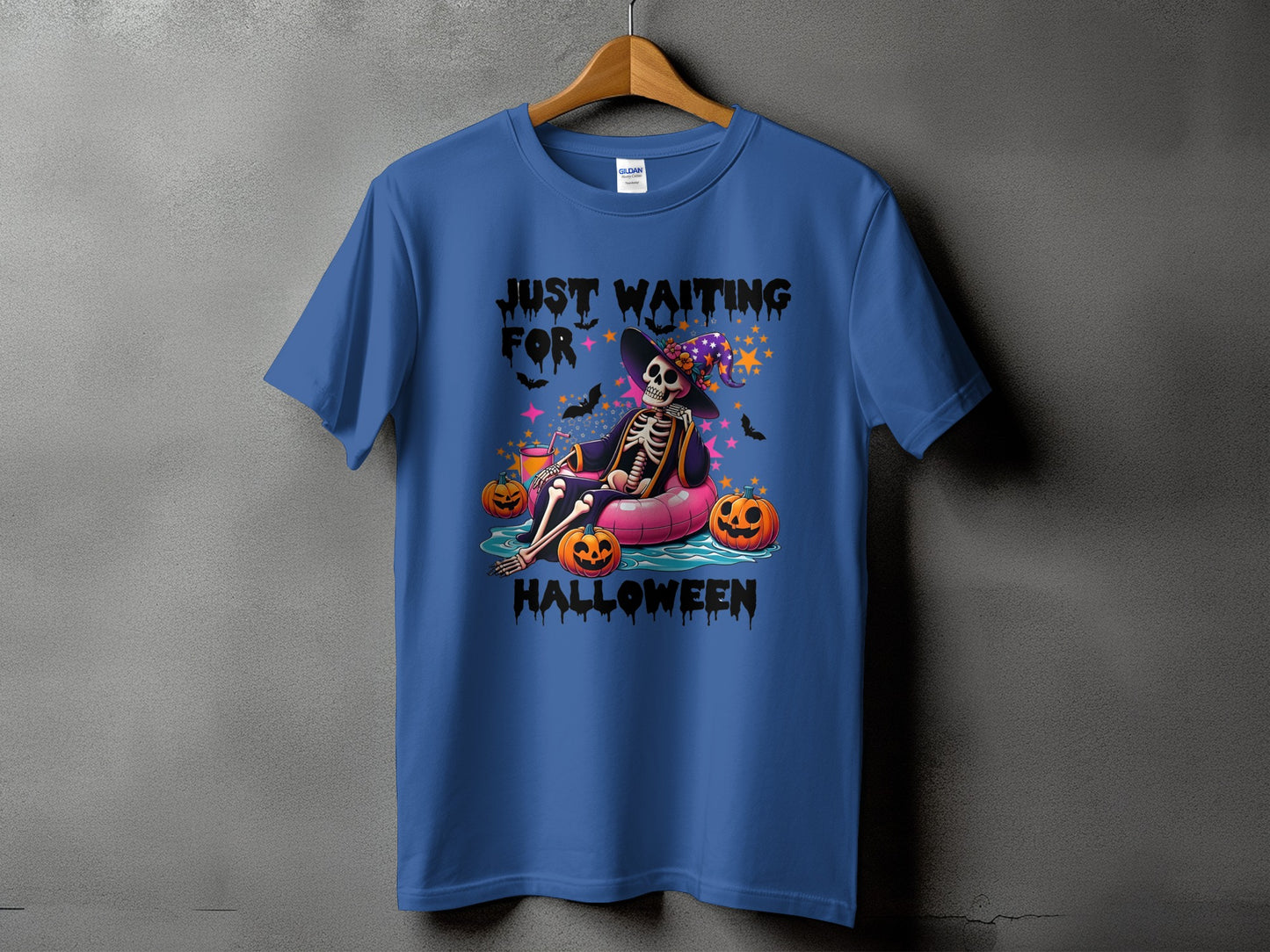 Skeleton in a witch hat lounging on an inflatable, with pumpkins and bats, saying "Just Waiting For Halloween".