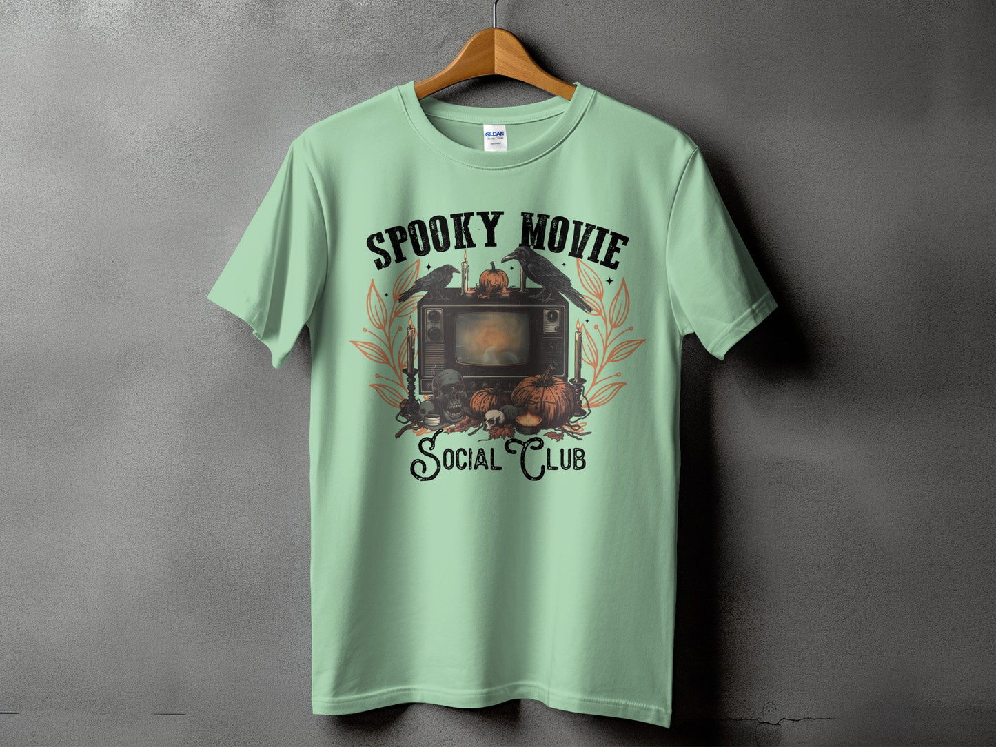 Light green t-shirt with "Spooky Movie Social Club" graphic featuring vintage TV and Halloween motifs.