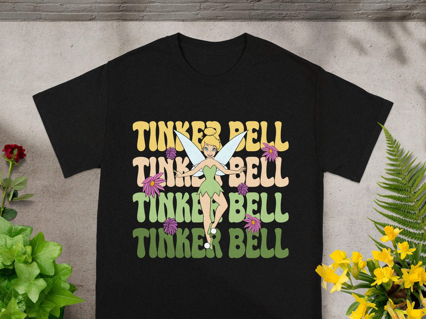 Black t-shirt with "Tinker Bell" in colorful, layered text and fairy illustrations.