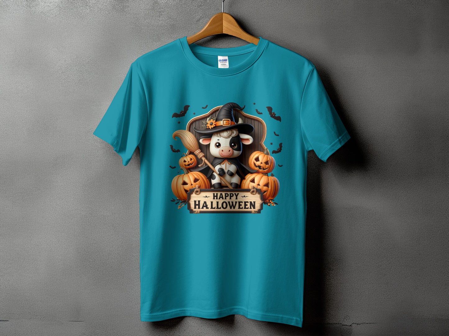 Teal t-shirt with cartoon cow in witch hat holding broom, surrounded by pumpkins and bats.
