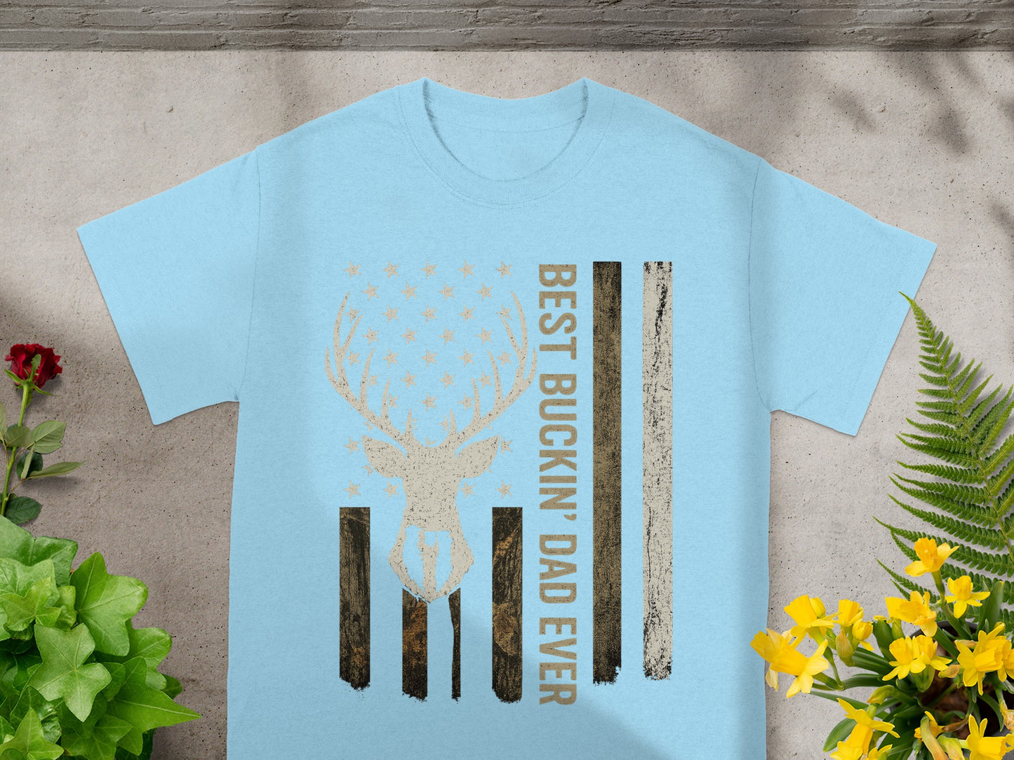 Light blue t-shirt with deer skull, stars, and American flag stripes. Text reads "BEST BUCKIN' DAD EVER".
