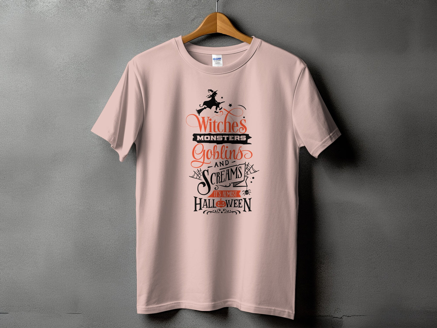 A dusty pink t-shirt hanging on a wooden hanger features black and orange Halloween-themed text and graphics.