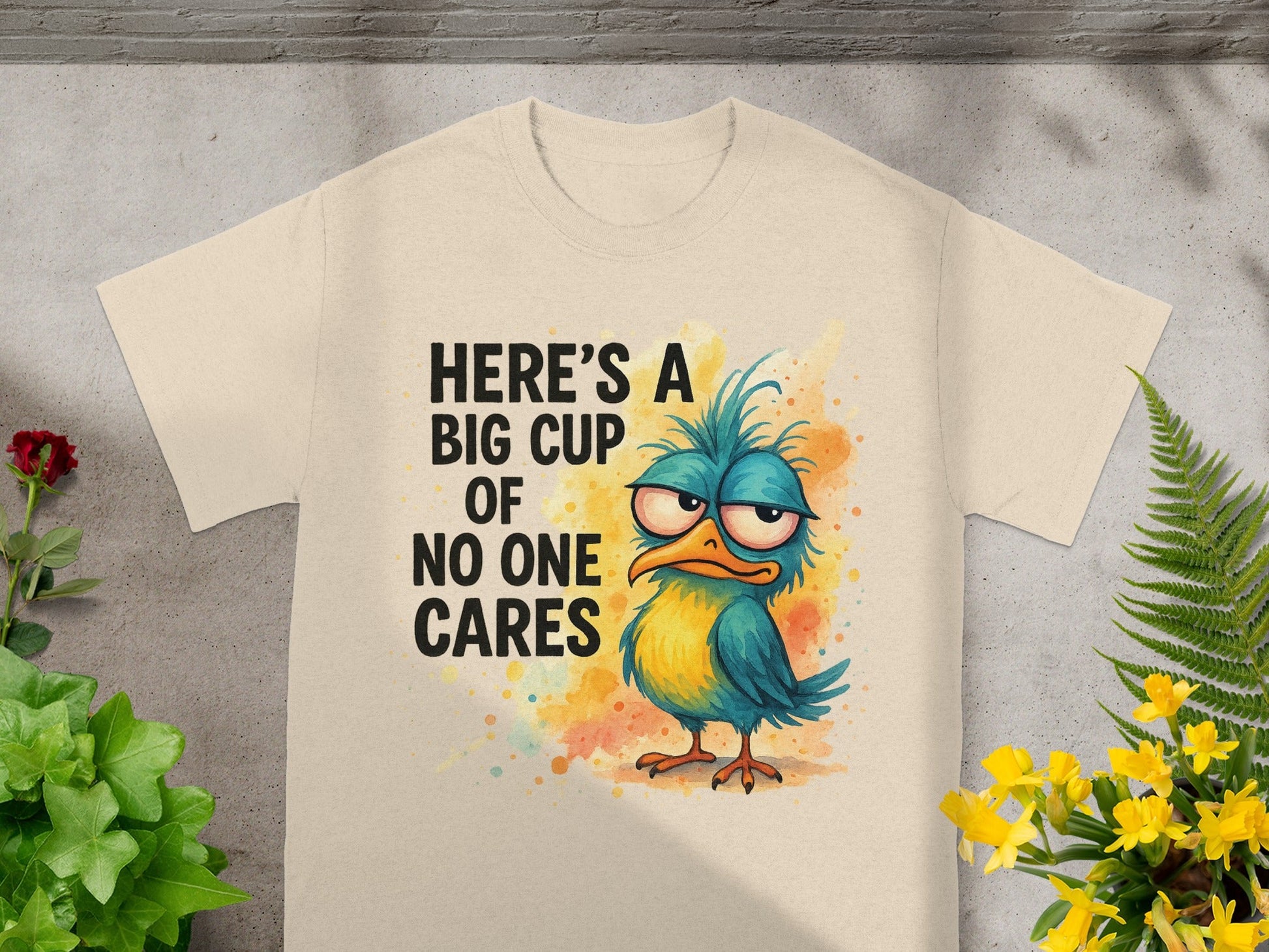Cream t-shirt with a grumpy blue and yellow cartoon bird and text "Here's a big cup of no one cares."