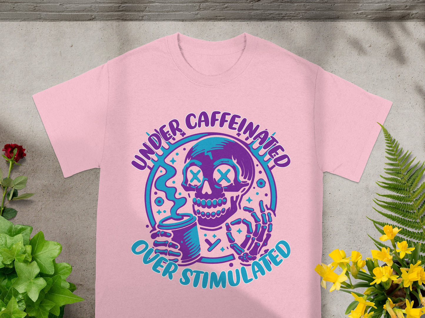 Pink t-shirt with a graphic of a skeleton holding a coffee cup, text "UNDER CAFFEINATED OVER STIMULATED".