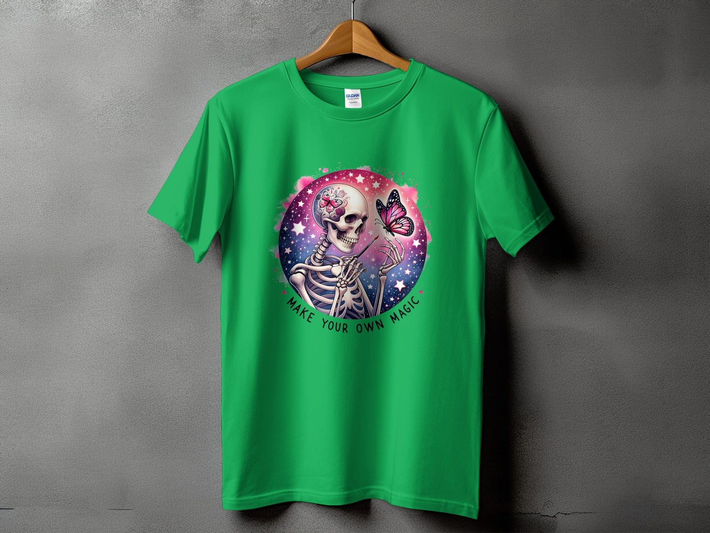 Green t-shirt with skeleton painting butterfly on cosmic background and "Make Your Own Magic" text.