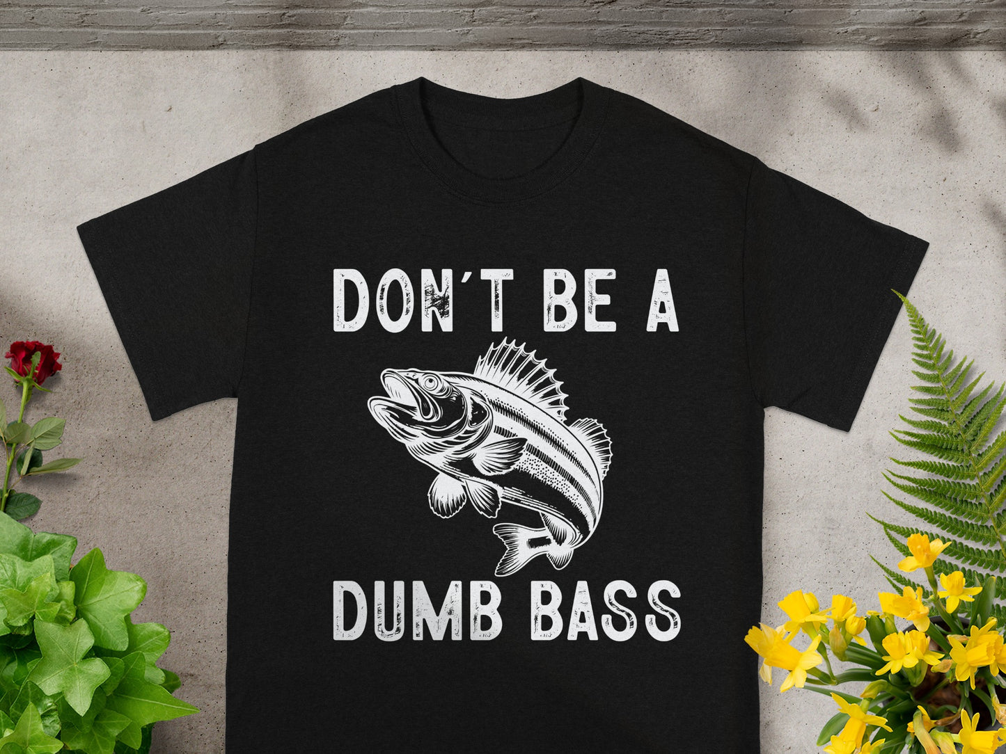 Black t-shirt with white text "DON'T BE A" above a bass fish illustration, and "DUMB BASS" below.