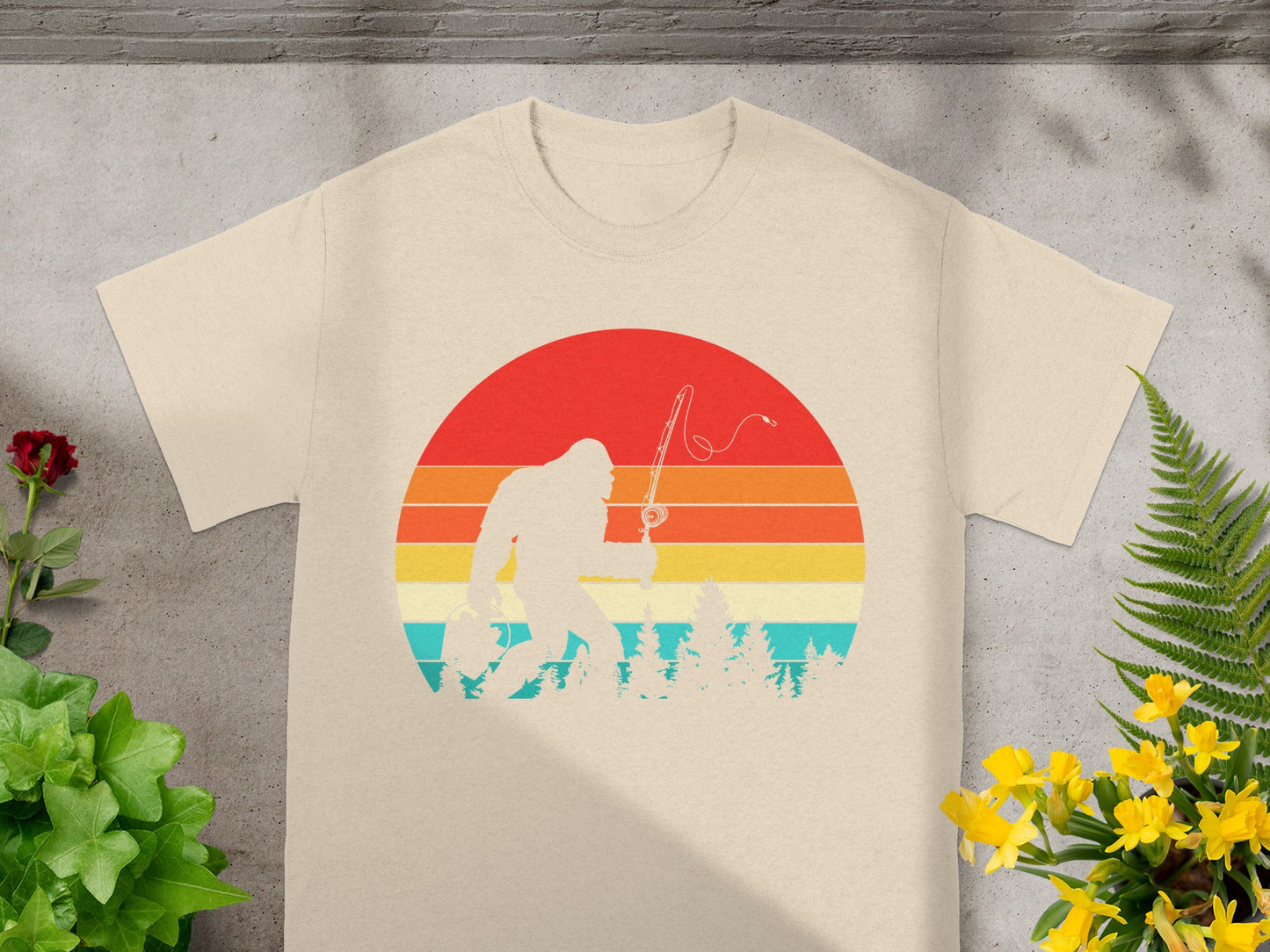 Bigfoot silhouette fishing with a sunset and trees design on a tan t-shirt.