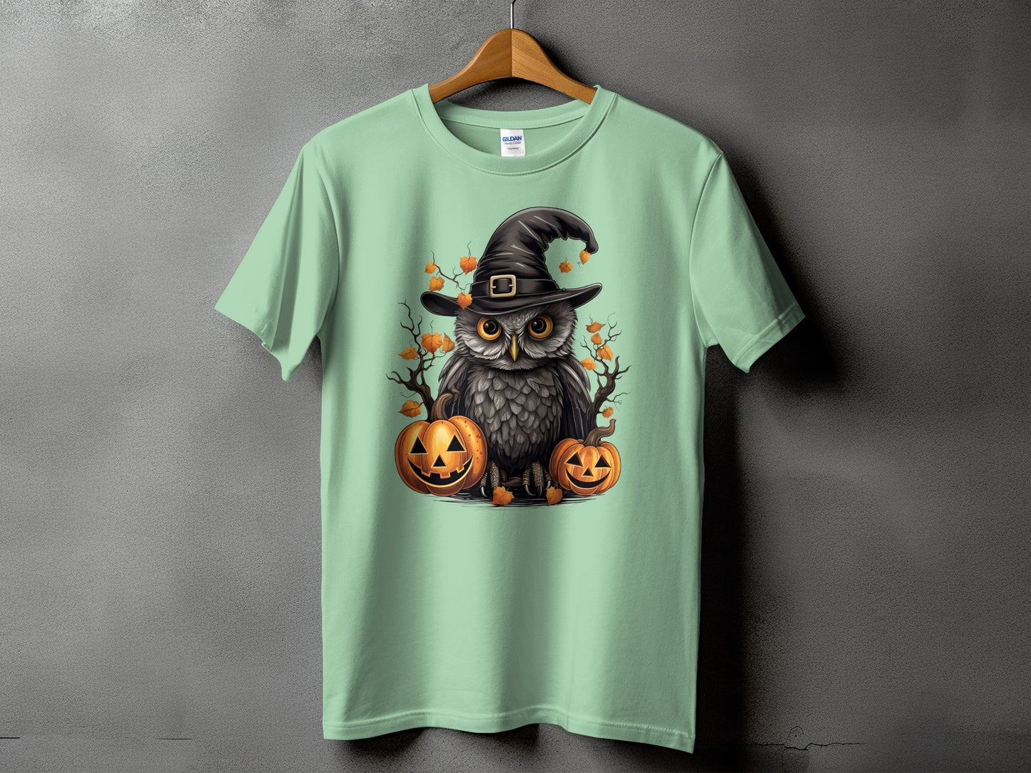 Owl in witch hat with pumpkins and autumn leaves graphic on a light green t-shirt.