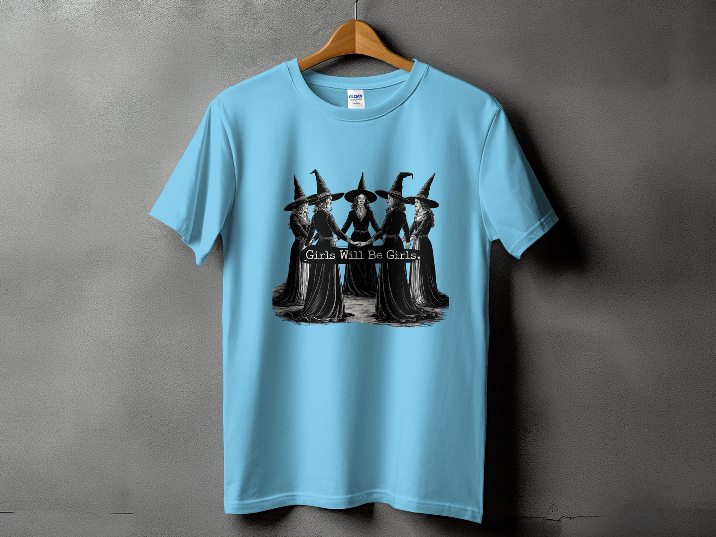 Light blue t-shirt featuring a graphic of five women in witch hats and text "Girls Will Be Girls."