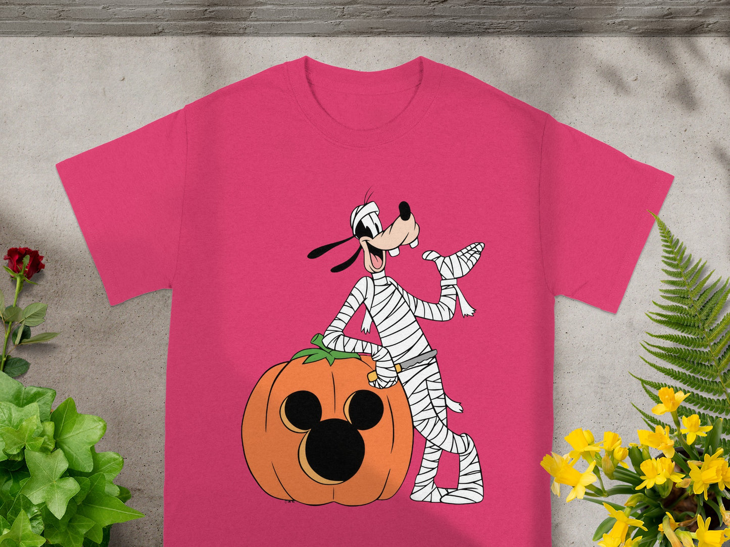 Goofy in mummy costume leans on a Mickey Mouse jack-o'-lantern on a pink t-shirt.