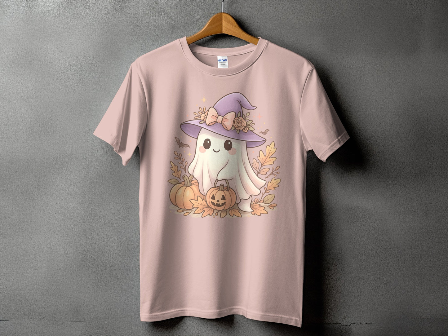 Pink t-shirt with a smiling ghost wearing a witch hat, holding a pumpkin basket, surrounded by pumpkins and leaves.
