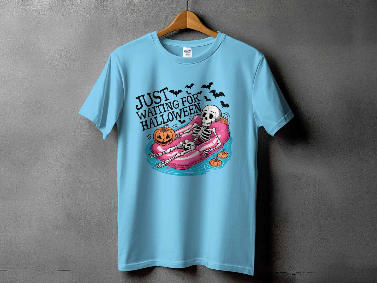 Skeleton floating on pink tube with pumpkins and bats, text "JUST WAITING FOR HALLOWEEN".