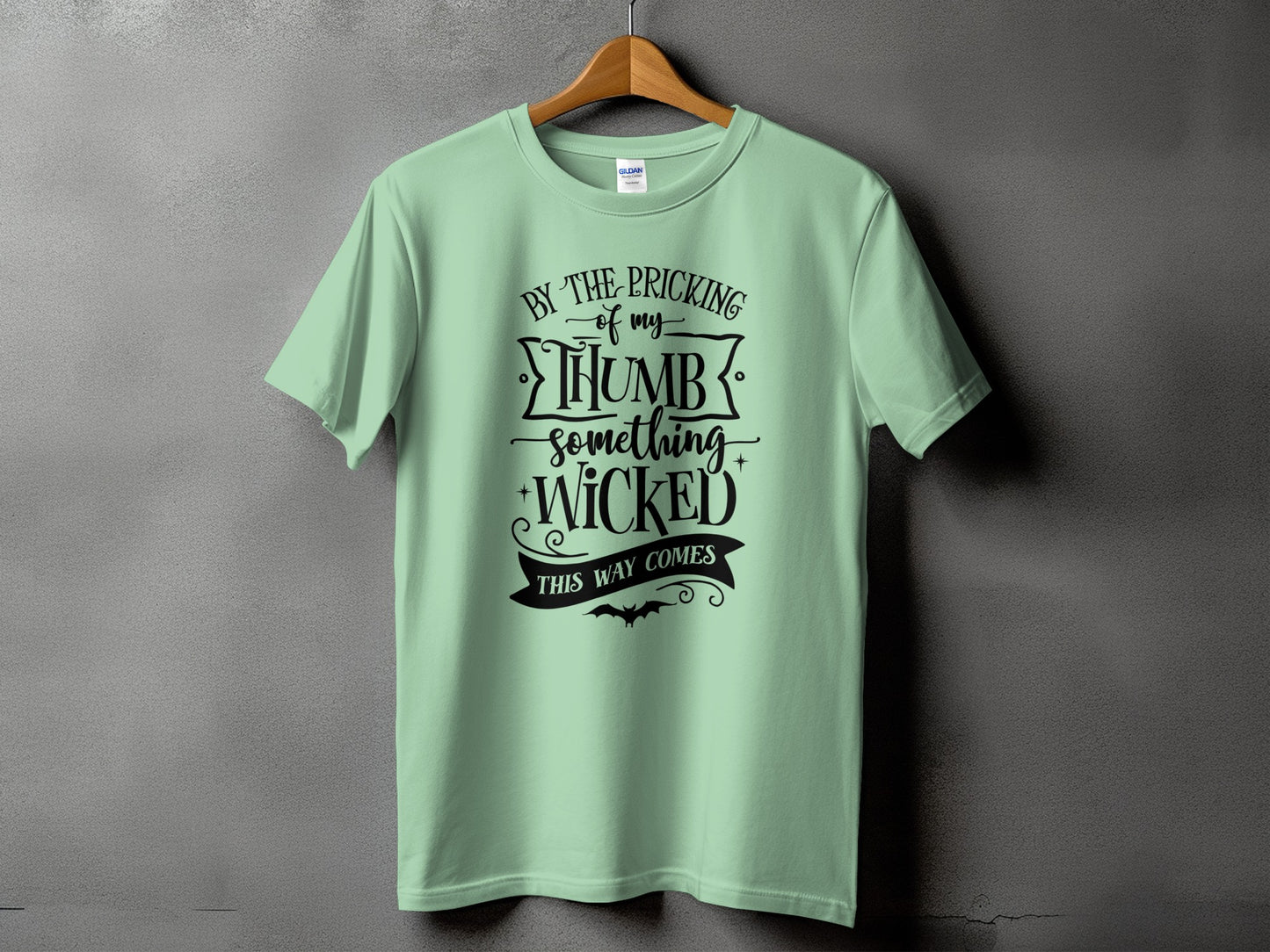 Light green t-shirt with black text reading "By the pricking of my thumb something wicked this way comes."