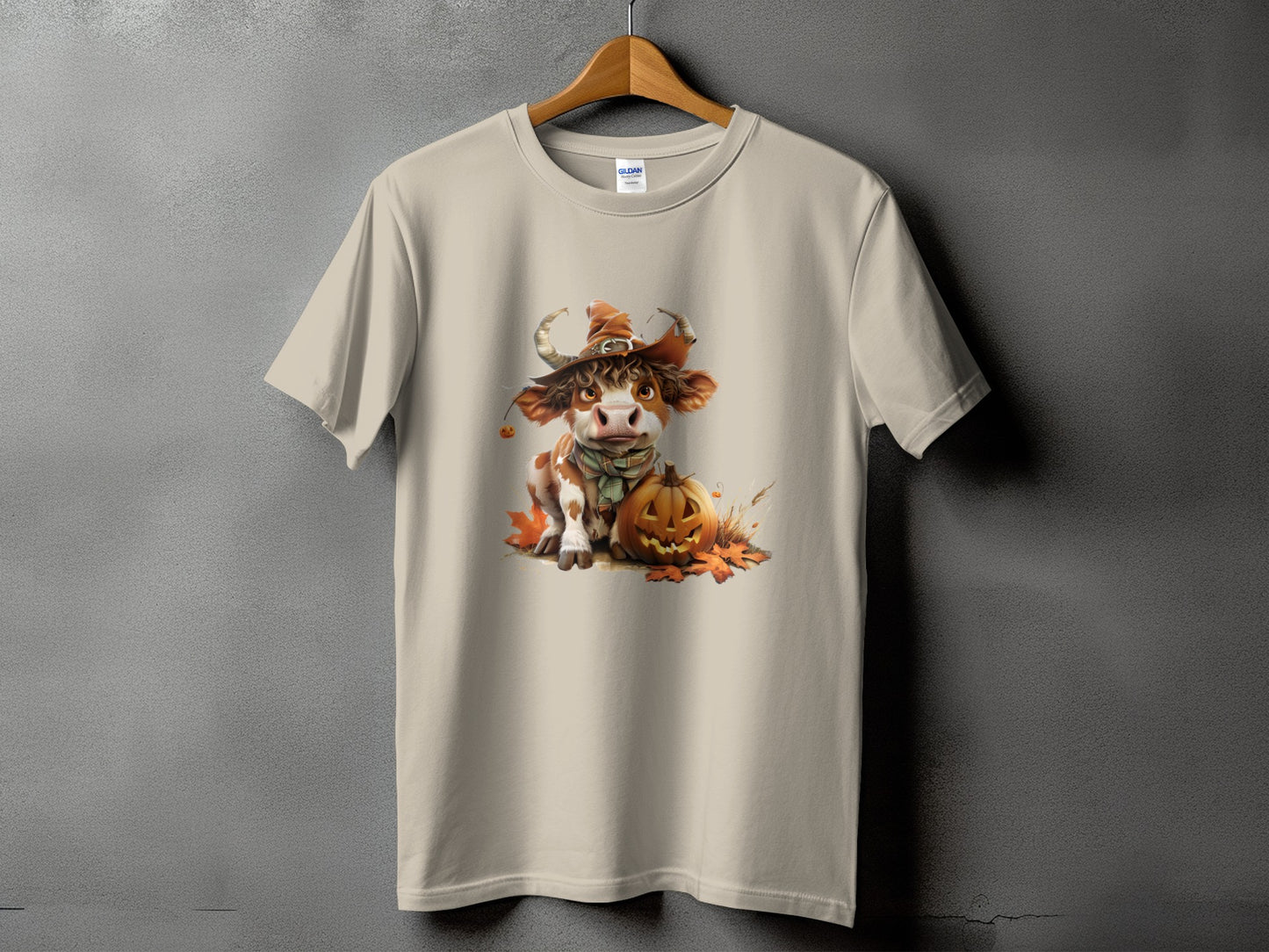 Cartoon cow wearing witch hat and scarf with pumpkin and autumn leaves on a tan t-shirt.