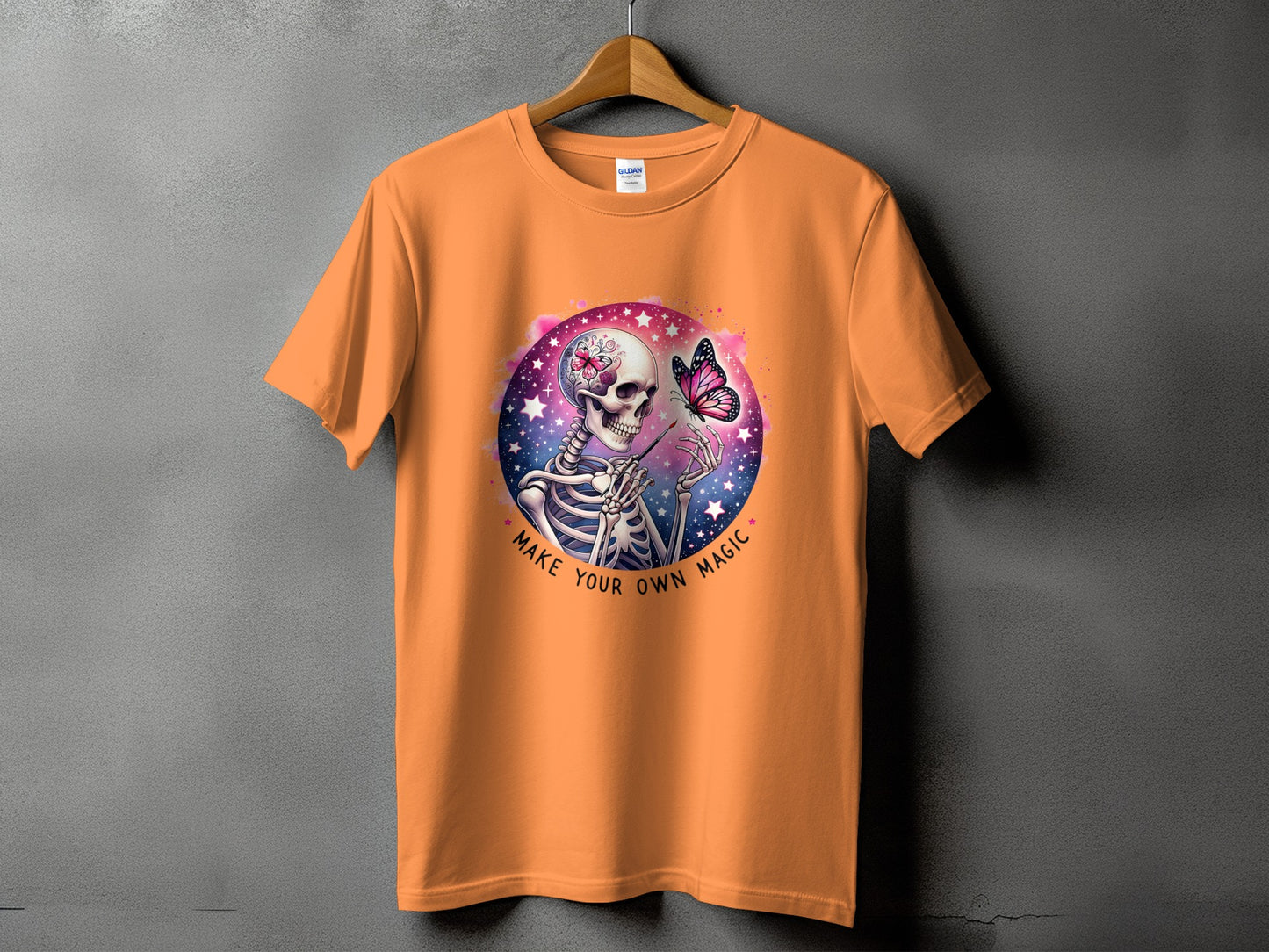 Orange t-shirt with skeleton painting a butterfly with the words "Make Your Own Magic".