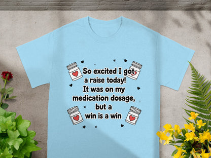 Light blue t-shirt with hearts and medicine bottles; text reads "So excited I got a raise today! It was on my medication dosage, but a win is a win."