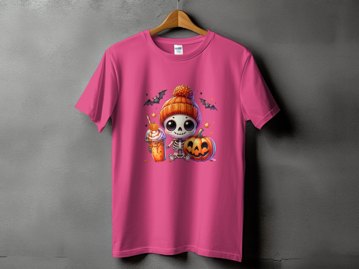 Pink t-shirt with a cartoon skeleton wearing a pumpkin hat, holding a drink, and sitting next to a pumpkin.
