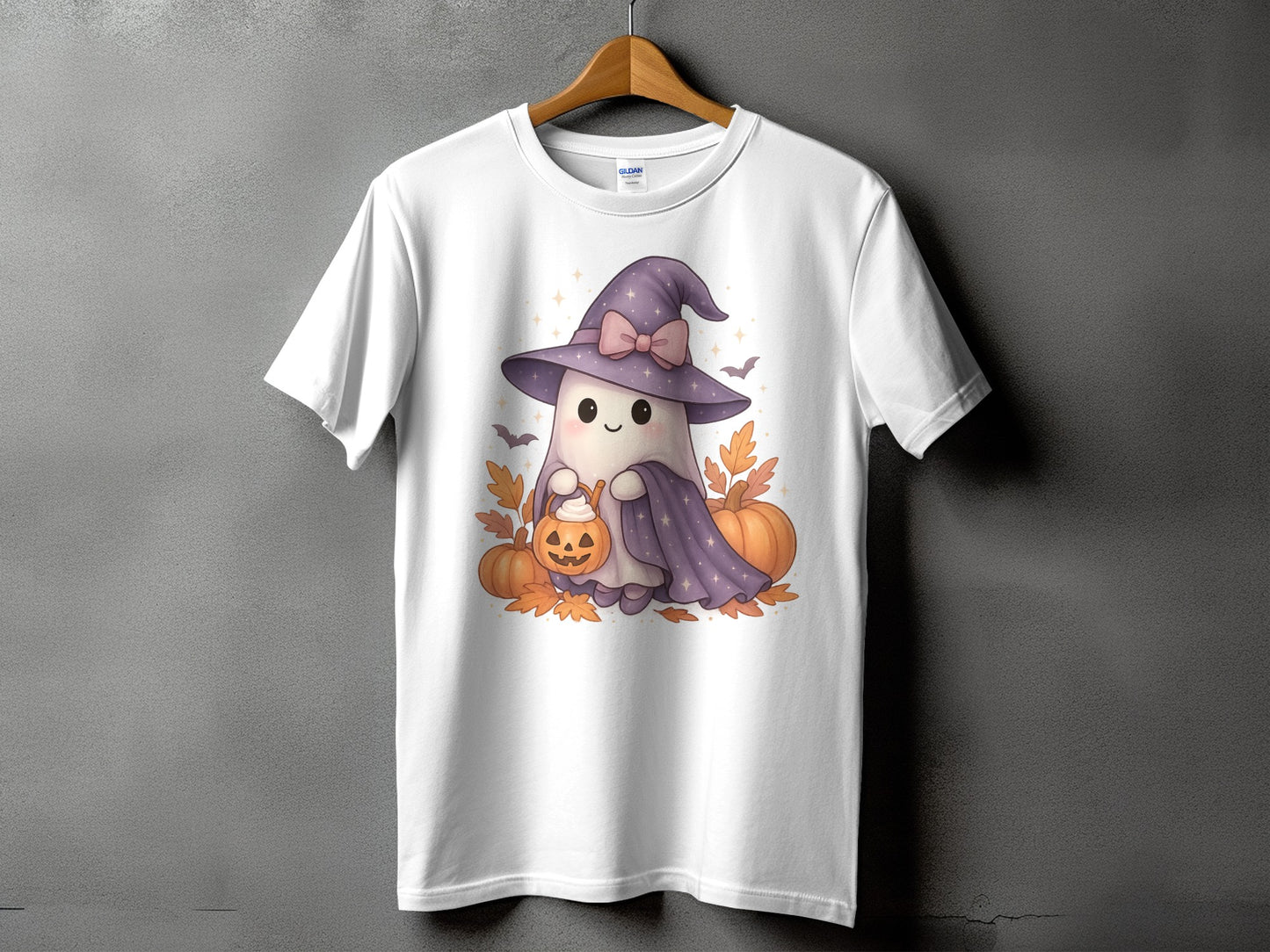 White t-shirt with cartoon ghost in witch hat holding pumpkin, surrounded by pumpkins and leaves.