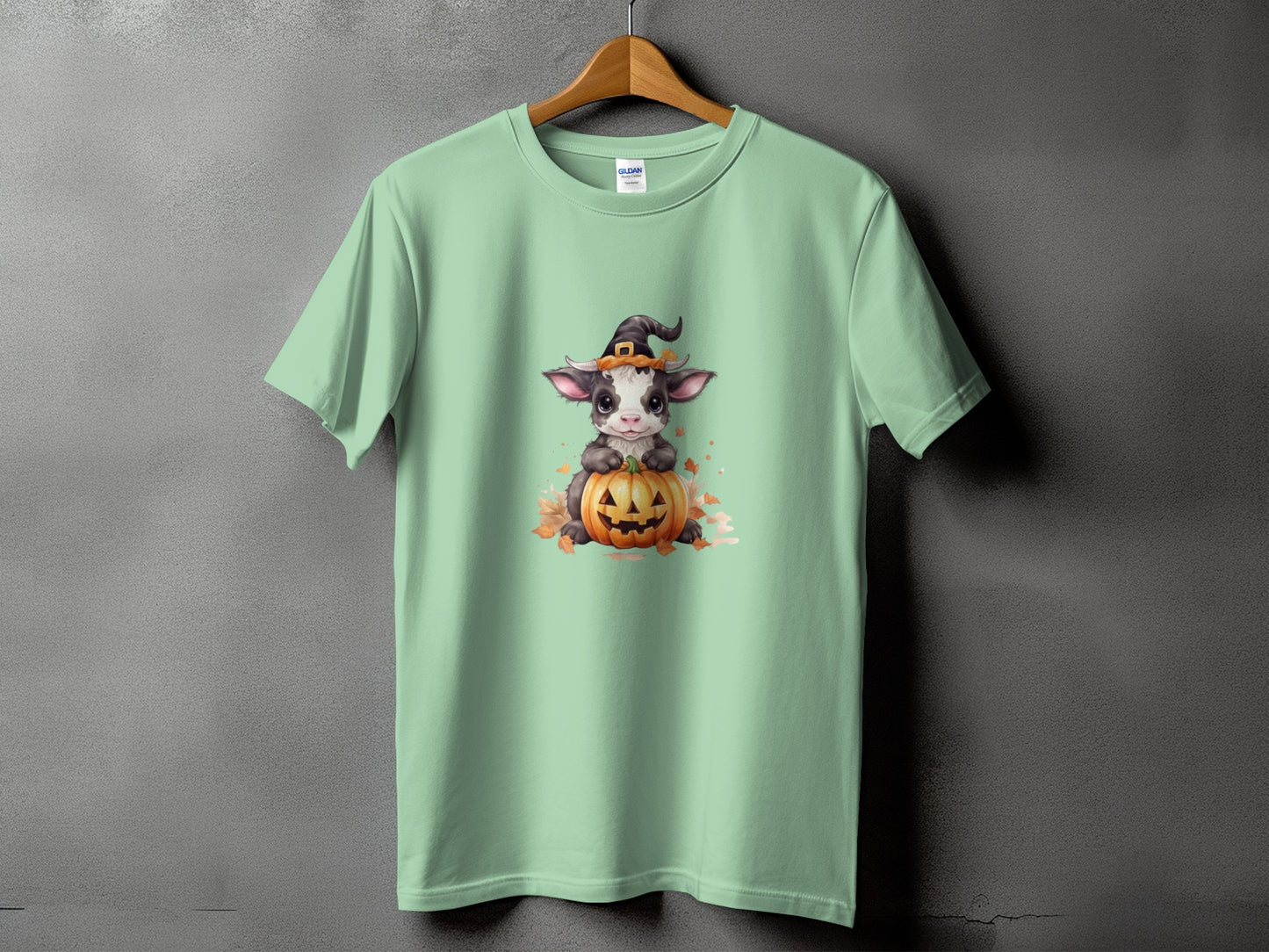 Light green t-shirt with a cartoon cow wearing a witch hat and sitting on a pumpkin.