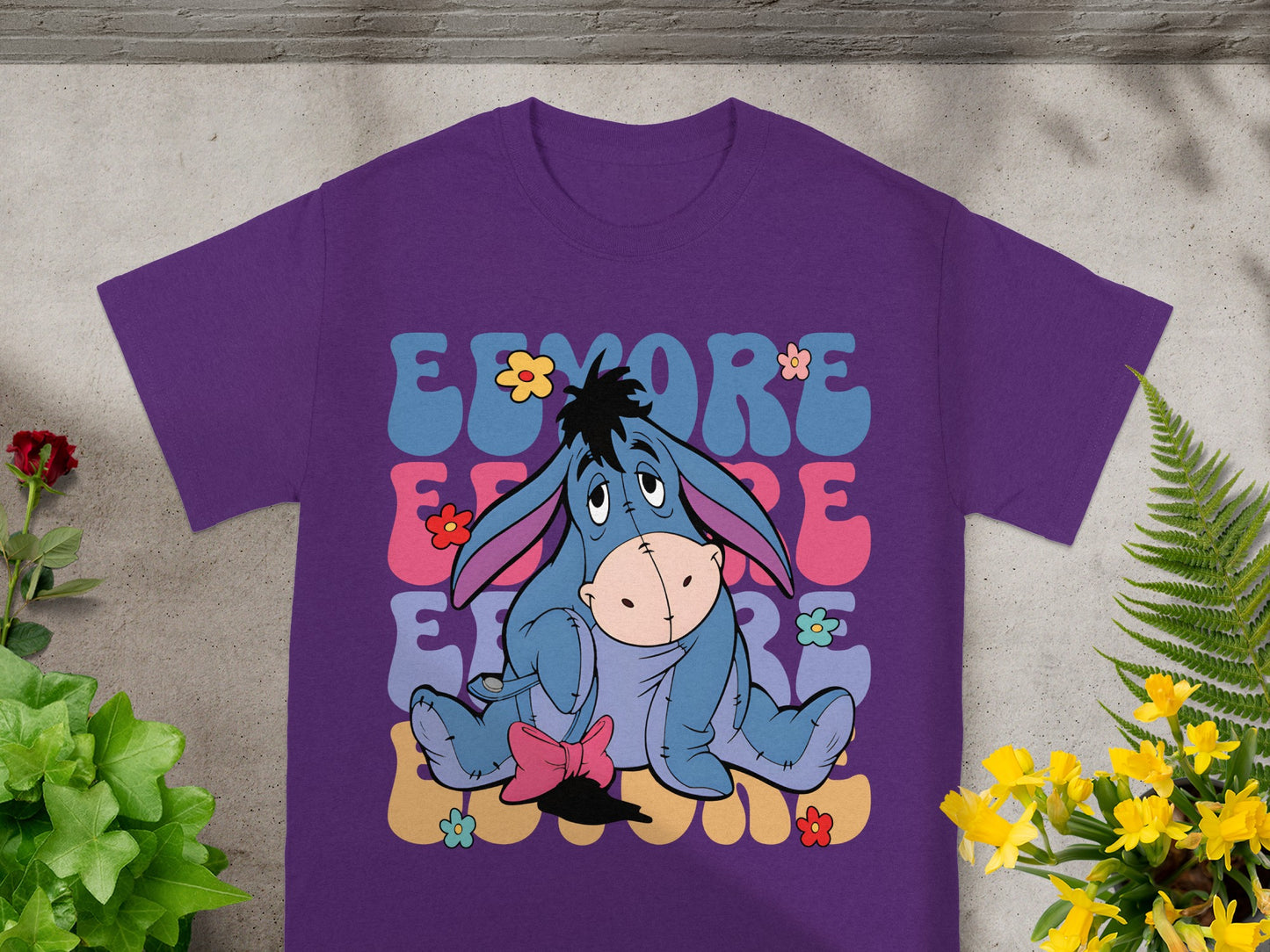 Purple t-shirt with Eeyore sitting among colorful flowers and text.