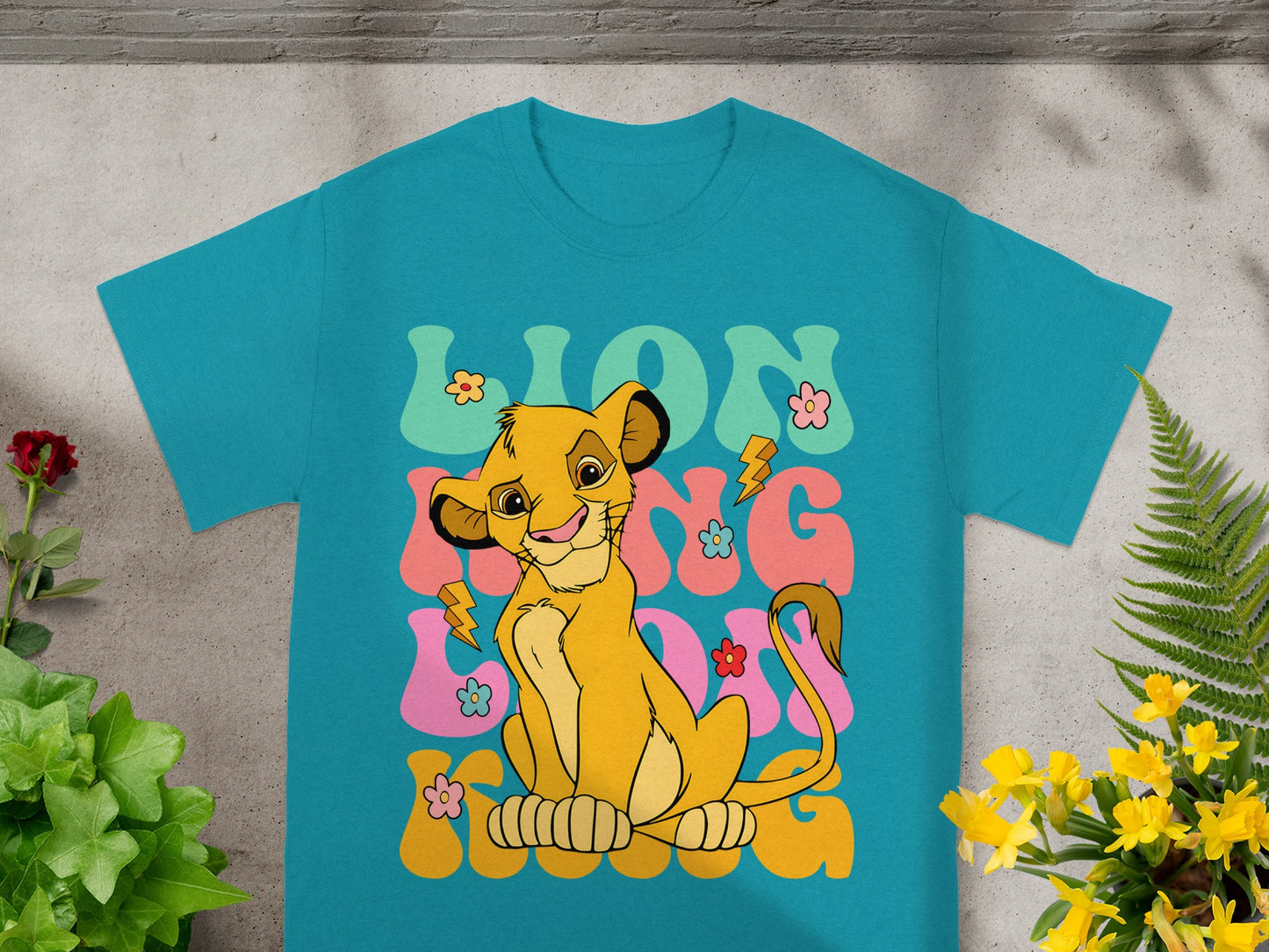 Simba from The Lion King sits smiling on a teal t-shirt with colorful text and flowers.