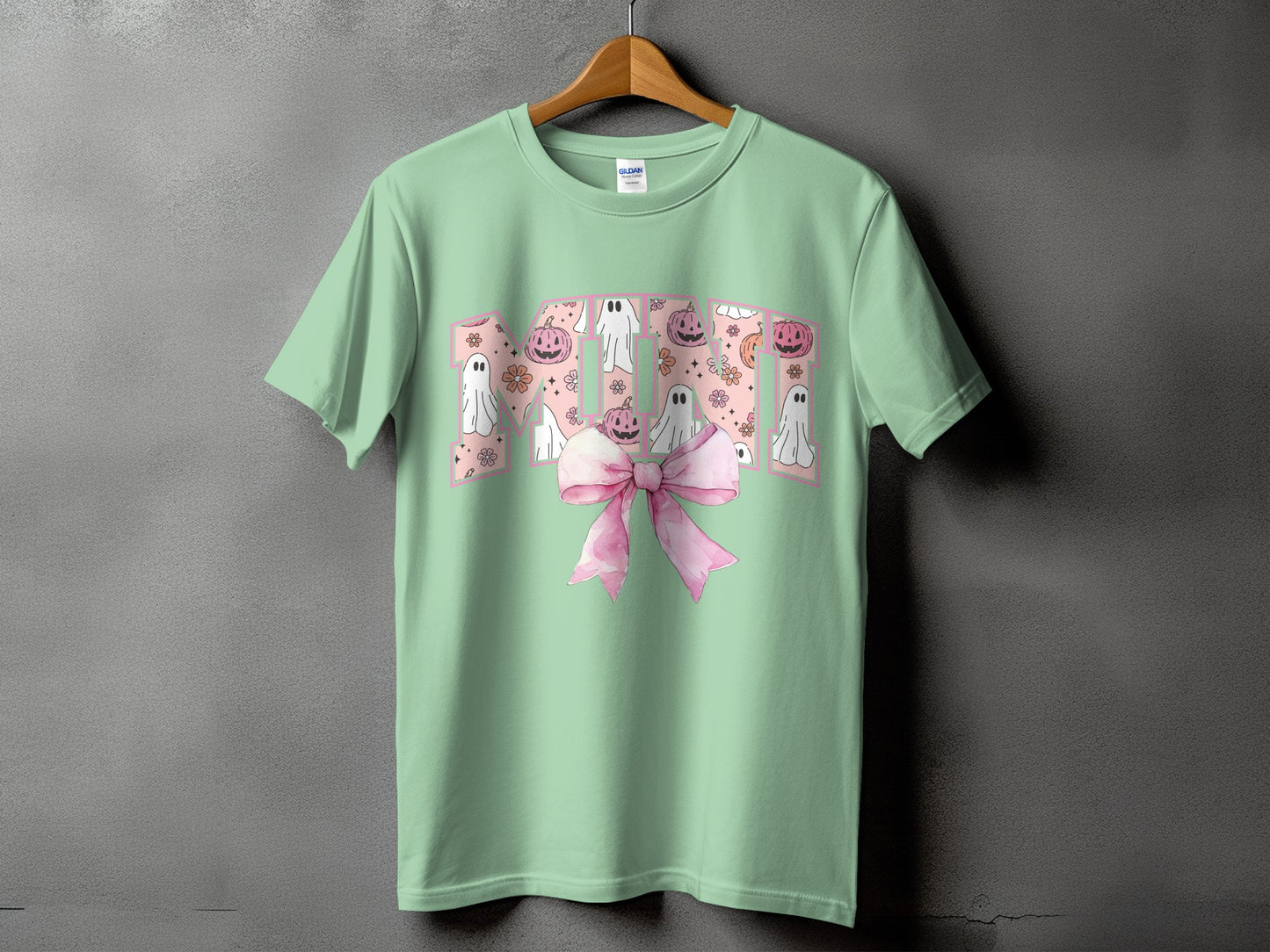 Green t-shirt with pink "MOM" text featuring ghosts, pumpkins, and flowers, and a pink bow.